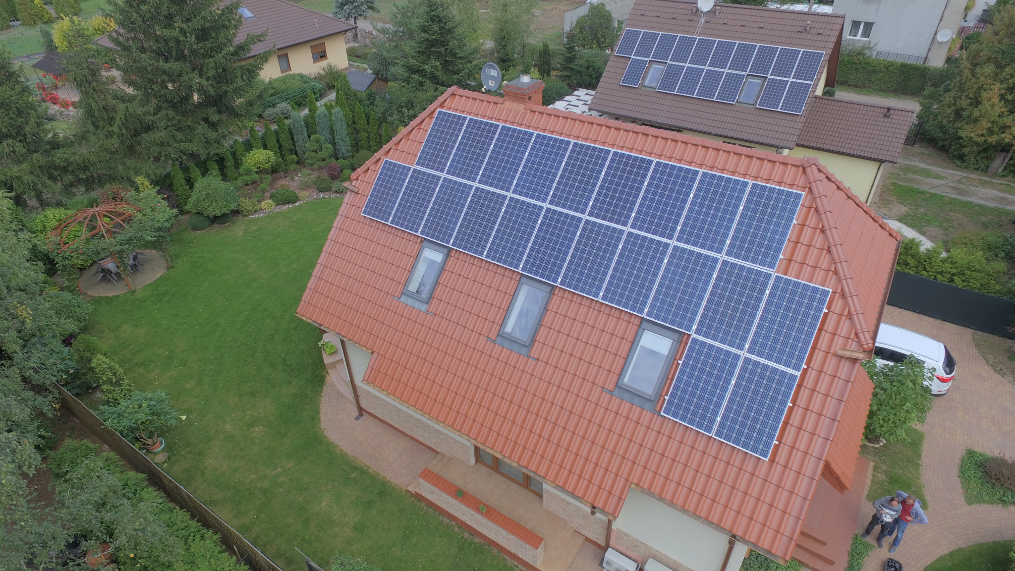 Widok z drona na dom jednorodzinny w Opławcu z panelami słonecznymi na dachu z czerwonej dachówki, obok zaparkowany biały samochód i zadbany ogród z altaną.