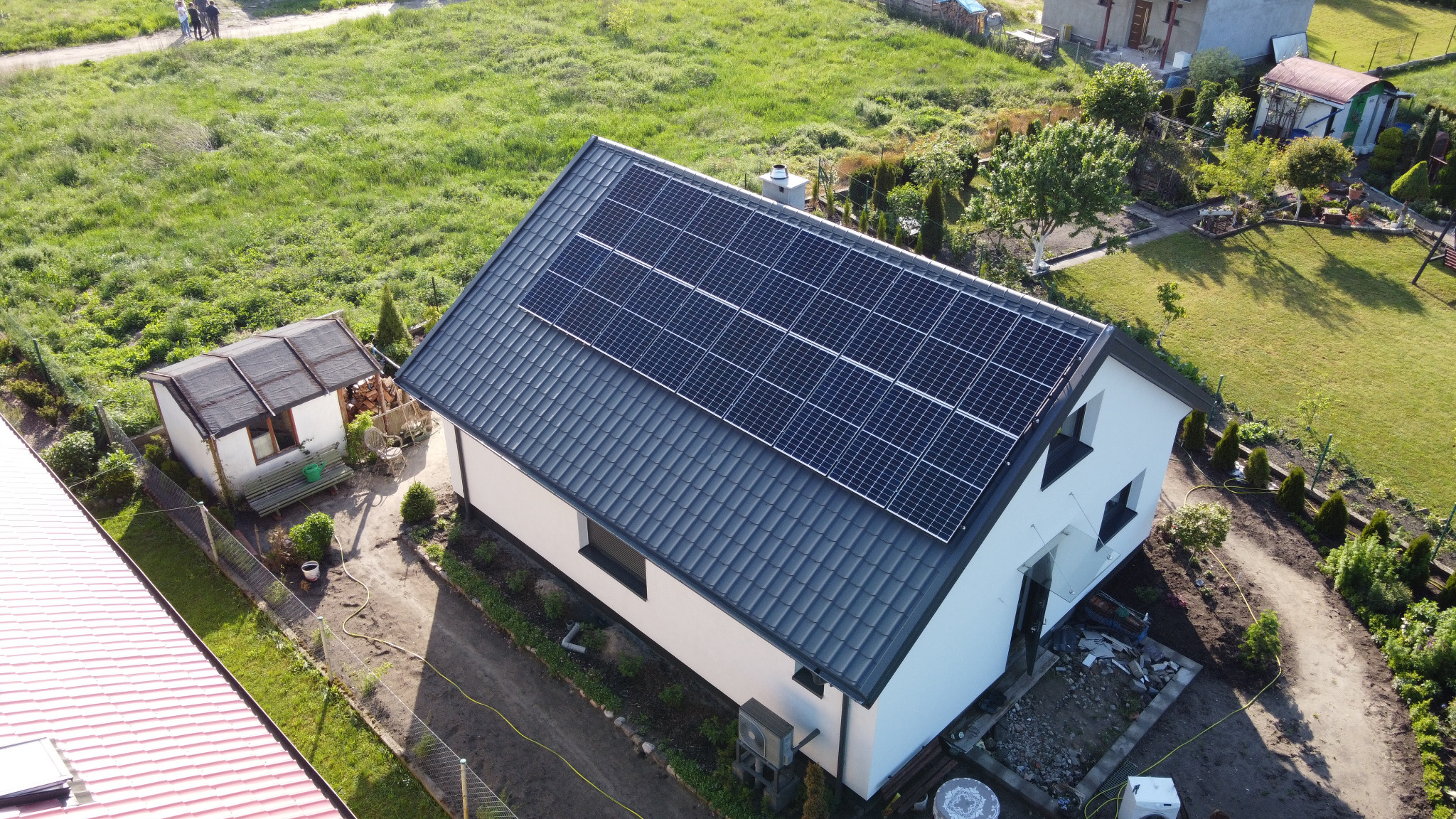 Widok z drona na biały dom z szarą dachówką i panelami słonecznymi na dachu, otoczony zielonym ogrodem i dodatkową zabudową.
