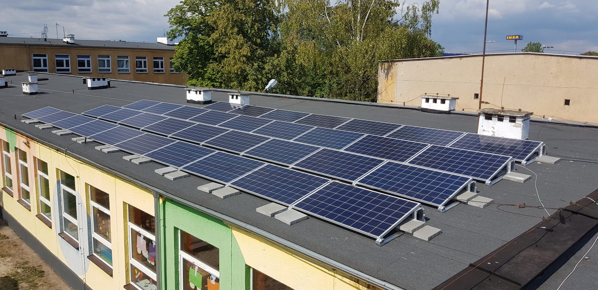 Panele słoneczne zamontowane na płaskim dachu budynku z kominami, widok z góry, w tle budynek IKEA.