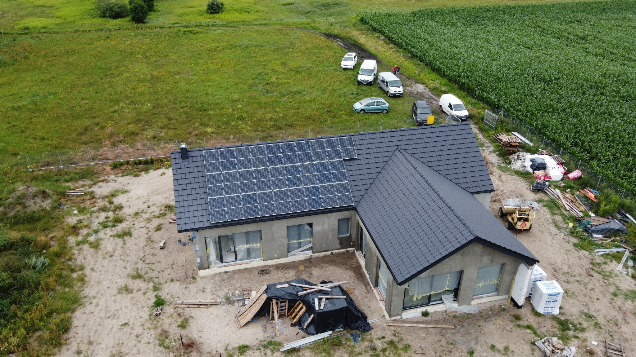 Widok z góry na dom jednorodzinny z zamontowanymi panelami słonecznymi na dachu, w tle pole kukurydzy i zaparkowane samochody, teren wokół domu w trakcie prac wykończeniowych.