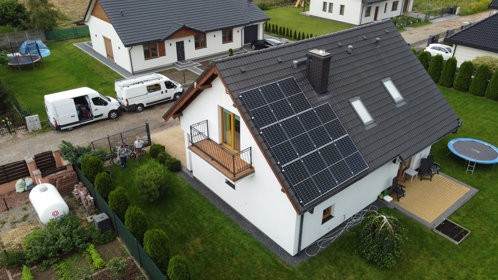 Widok z góry na dom z panelami słonecznymi na dachu, dwa białe samochody dostawcze zaparkowane obok, instalatorzy przy ogrodzeniu, zielony trawnik i zadbany ogród.