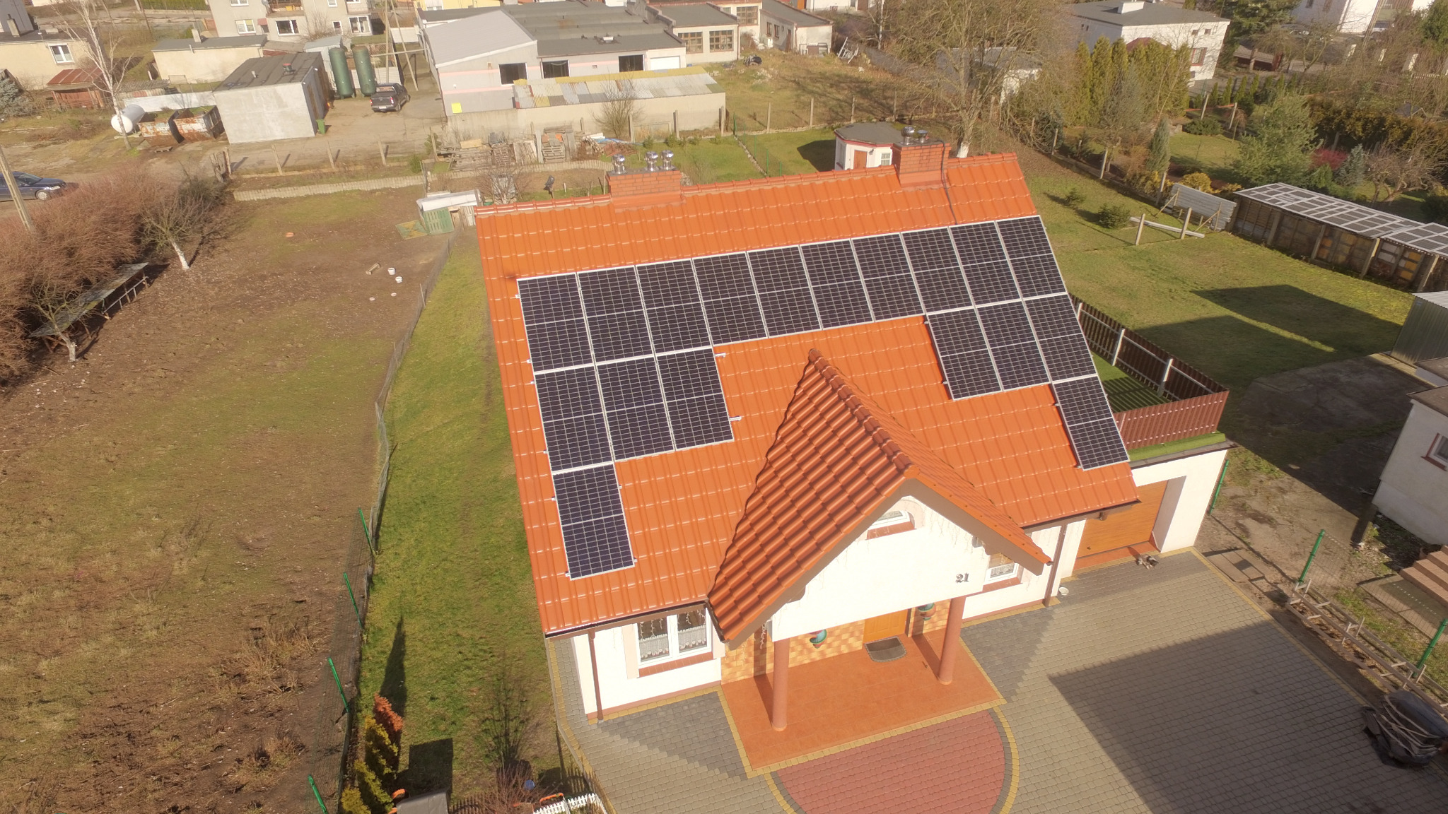 Widok z drona na dom jednorodzinny z pomarańczową dachówką i panelami słonecznymi, na posesji rosną drzewa, a w tle widać inne budynki.