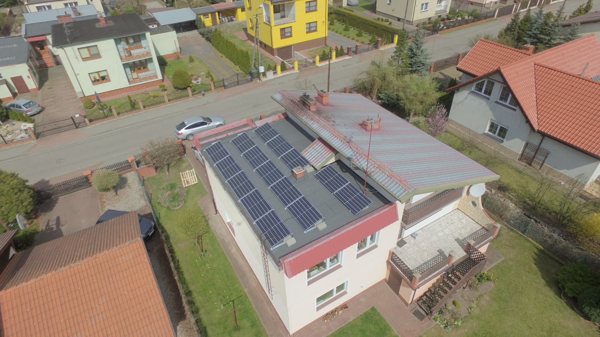 Widok z lotu ptaka na dom jednorodzinny z panelami słonecznymi na dachu, otoczony zielenią i zabudową sąsiedzką.