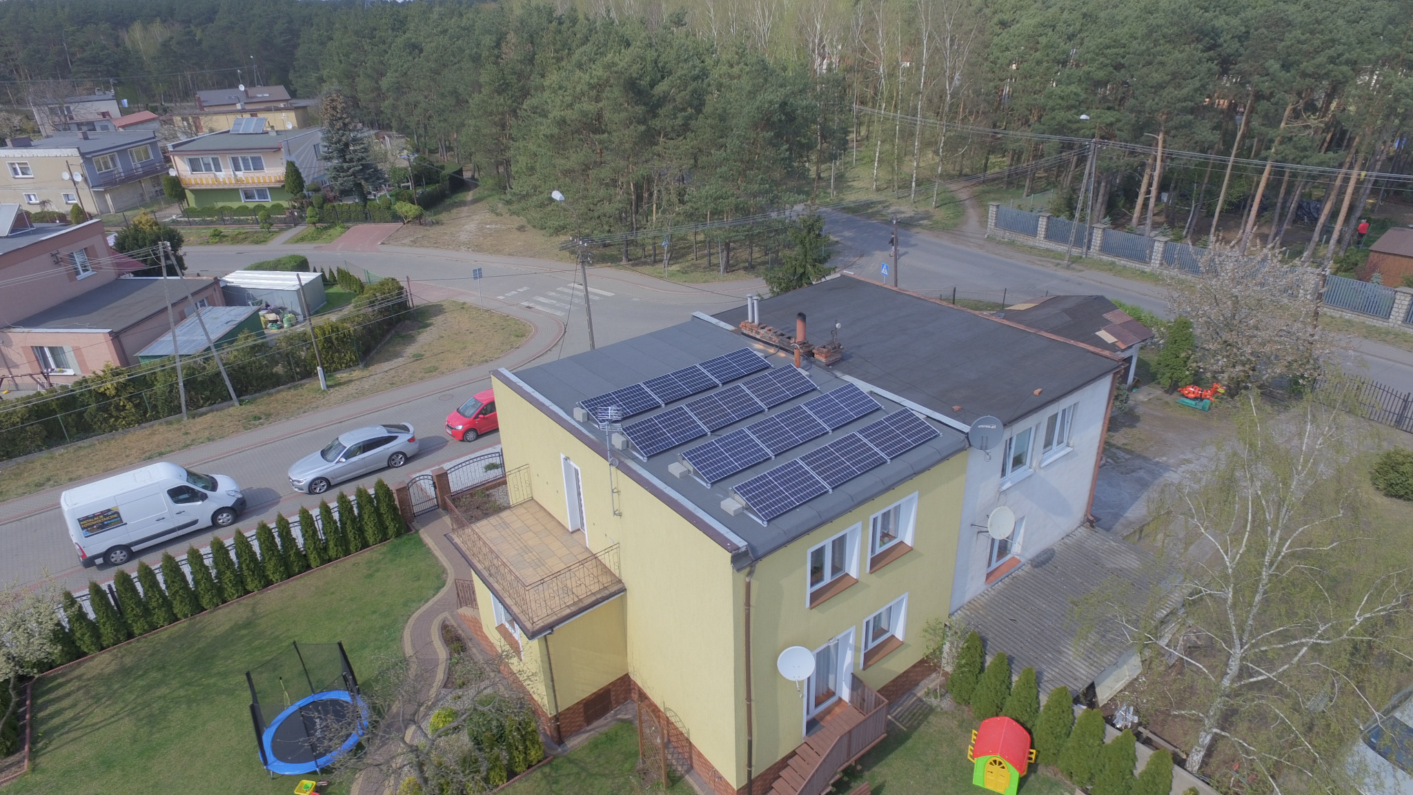 Widok z góry na dom z żółtą elewacją i zamontowanymi panelami słonecznymi na dachu, w tle ulica z samochodami i las.