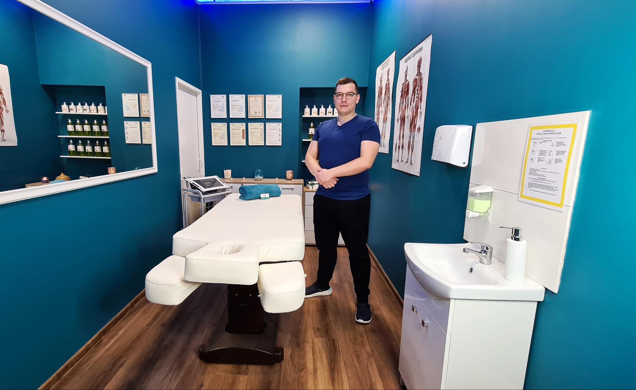 Wnętrze gabinetu z białym łóżkiem do zabiegów, umywalką i stojącym mężczyzną w okularach na tle ściany z certyfikatami i plakatem anatomicznym.