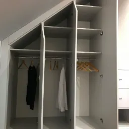Biała szafa wnękowa na poddaszu ze skosami, z otwartymi drzwiami, widoczne ubrania na wieszakach.