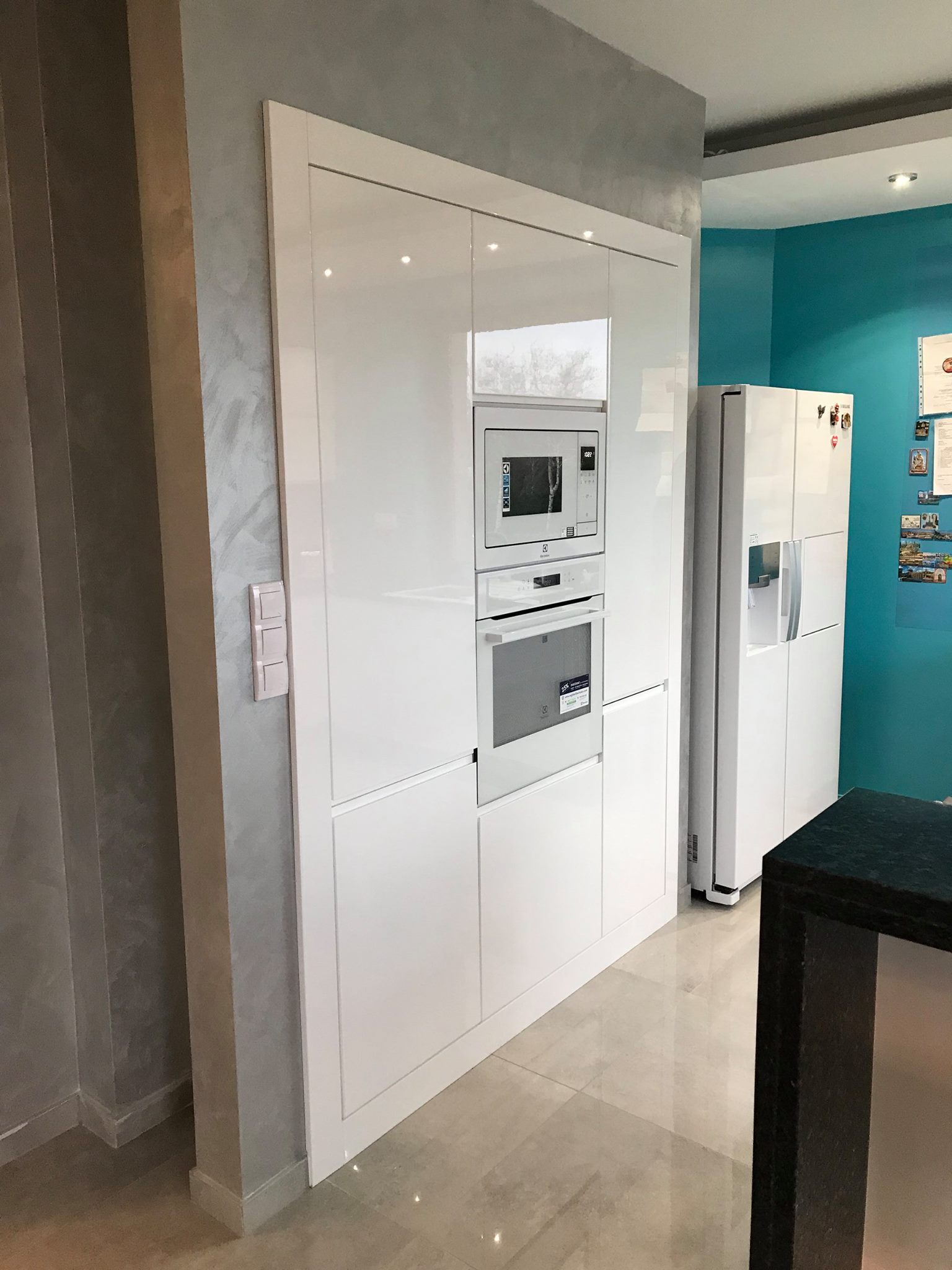 Biała, lakierowana zabudowa kuchenna z wbudowanym piekarnikiem i mikrofalówką Electrolux, sąsiadująca z lodówką side-by-side na tle turkusowej ściany.