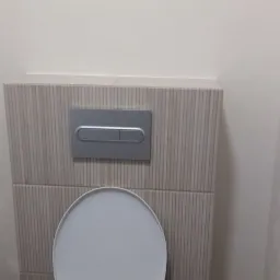 Zamontowana muszla klozetowa z podtynkową spłuczką obłożoną płytkami imitującymi drewno, na ścianie widoczny włącznik światła.