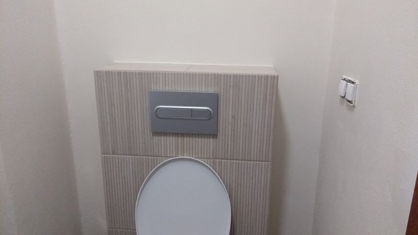 Zamontowana muszla klozetowa z podtynkową spłuczką obłożoną płytkami imitującymi drewno, na ścianie widoczny włącznik światła.