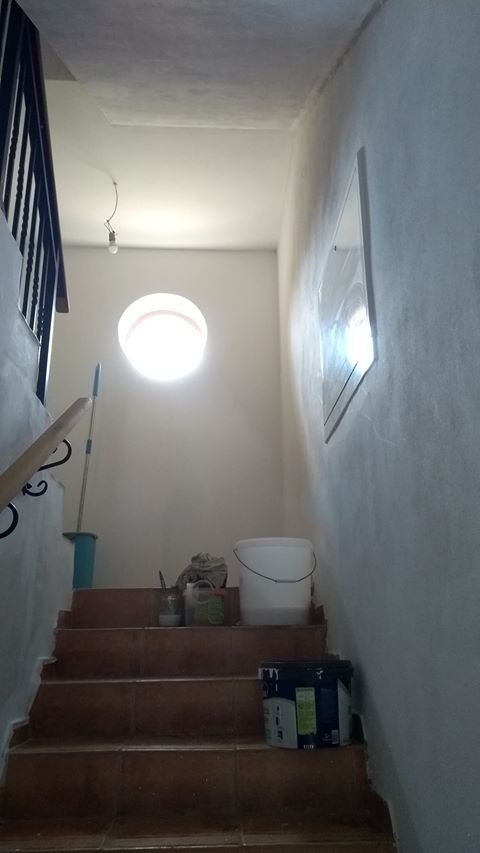 Klatka schodowa w trakcie odświeżania, widoczne narzędzia malarskie, wiadro z farbą i pędzel na schodach z terakoty, ściany w trakcie malowania na biało, okrągłe okno na ścianie.