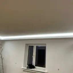 Instalacja oświetlenia LED w suficie podwieszanym przez montera stojącego na aluminiowej drabinie, widoczne okno i grzejnik.