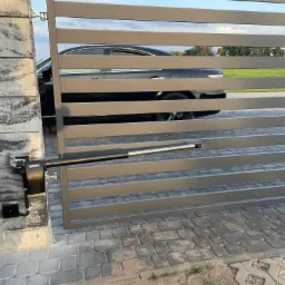 Szara, nowoczesna brama wjazdowa z automatycznym napędem FAAC, widoczny czarny samochód w tle, kostka brukowa na podjeździe.