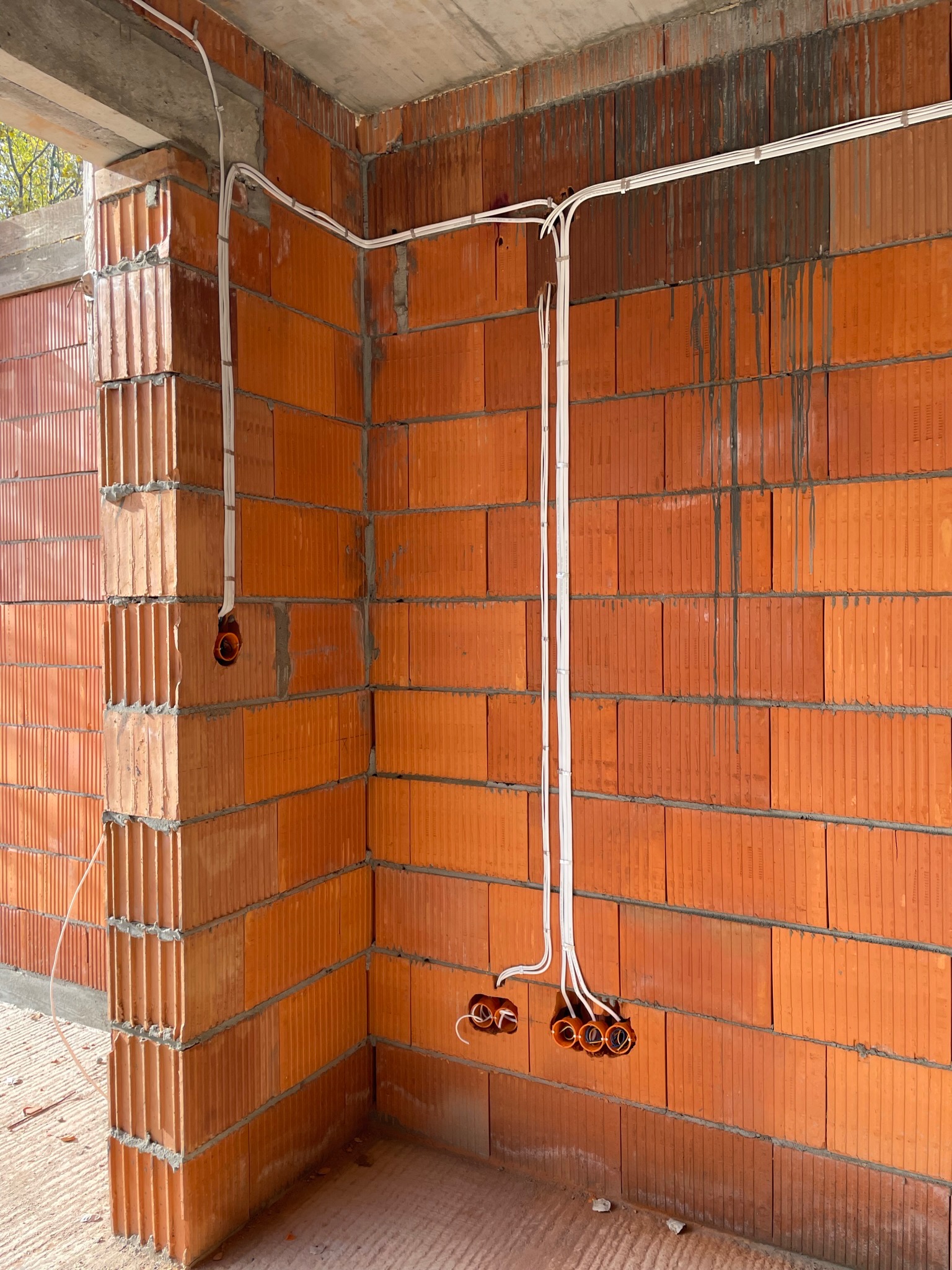 Surowy mur z czerwonej cegły z widocznymi przewodami elektrycznymi, pomarańczowe puszki instalacyjne, brak osprzętu, widok na wnętrze w trakcie budowy.