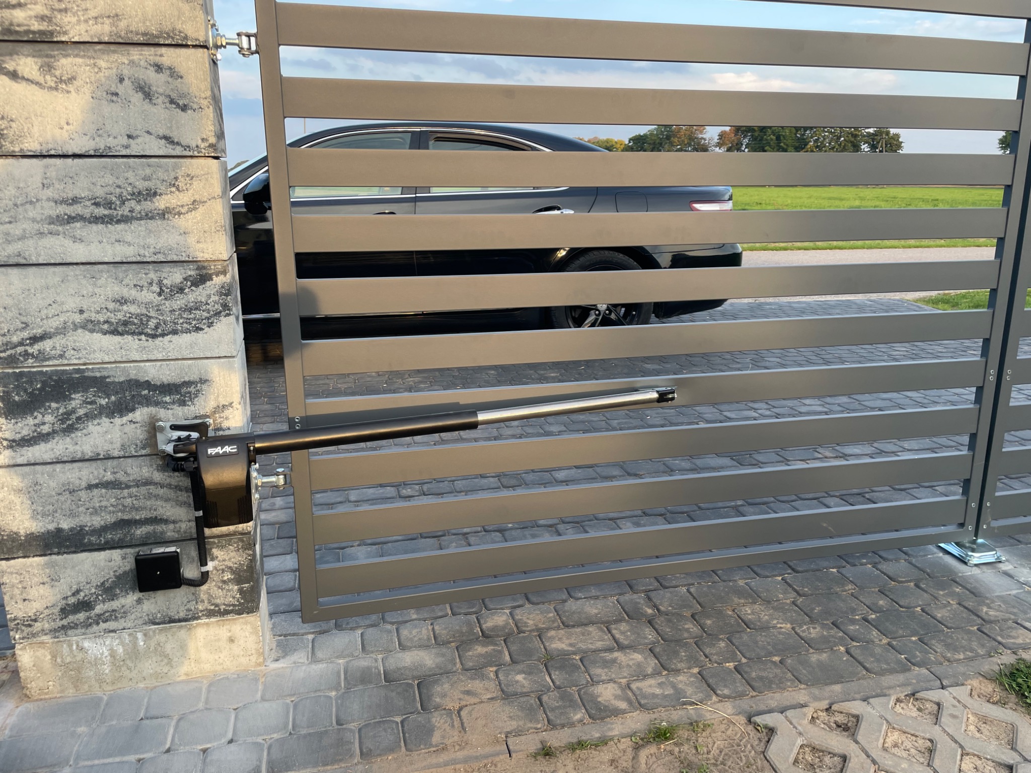 Szara, nowoczesna brama wjazdowa z automatycznym napędem FAAC, widoczny czarny samochód w tle, kostka brukowa na podjeździe.