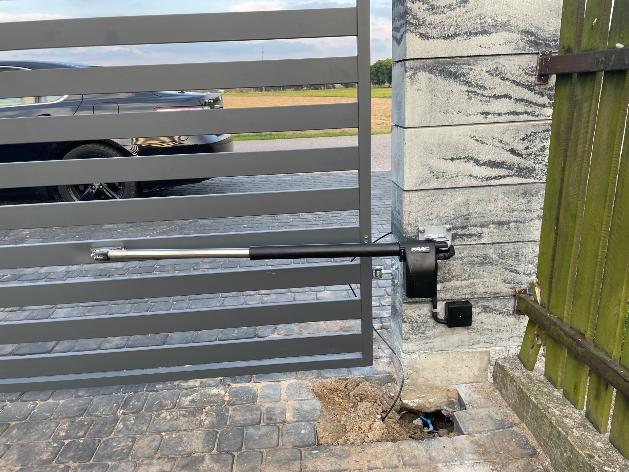 Szara brama przesuwna z zamontowaną automatyką FAAC, widoczne okablowanie w wykopanym otworze w kostce brukowej, w tle zaparkowany samochód.