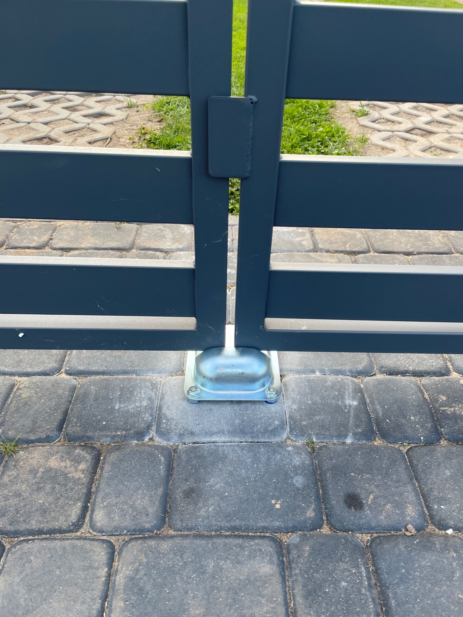 Szczegół mocowania szarej, metalowej bramy do kostki brukowej, widoczne śruby i podstawa mocująca.