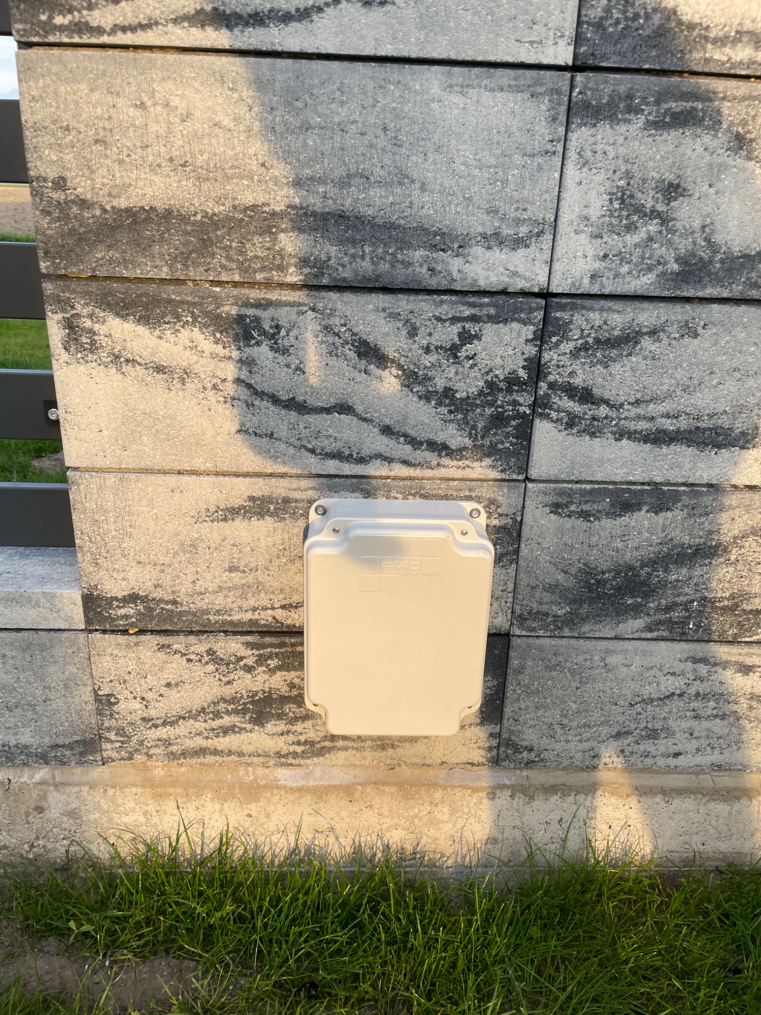 Szara skrzynka sterownicza FAAC zamontowana na betonowym słupku ogrodzenia z widoczną trawą u podstawy.