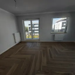 Nowoczesne wnętrze z parkietem ułożonym w jodełkę, widok na budynek mieszkalny przez balkonowe okno.