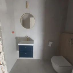 Wykończona łazienka z szarymi płytkami, wiszącą szafką z umywalką w kolorze granatowym z złotymi uchwytami, okrągłym lustrem w złotej ramie, lampą ścienną w kolorze złotym i podwieszaną toaletą.