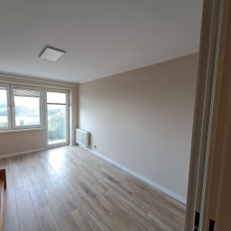 Aro Klimczak Arkadiusz - Wnętrze pustego pokoju z drewnopodobną podłogą z płytek, grzejnikiem pod oknem balkonowym i białymi ścianami, widok zza otwartych drzwi.