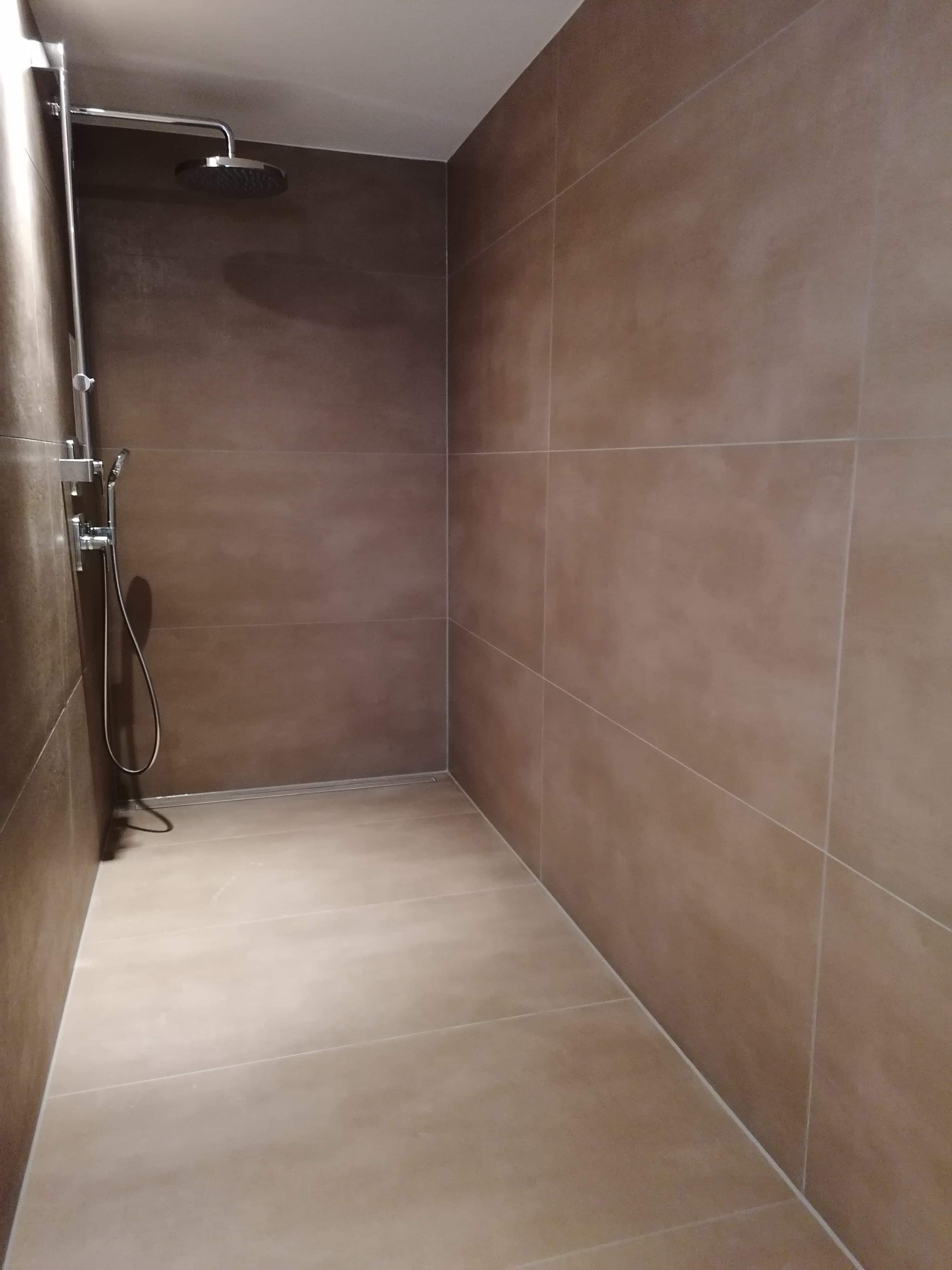 Wykończona kabina prysznicowa z brązowymi, matowymi płytkami na ścianach i podłodze, wyposażona w nowoczesny zestaw prysznicowy z deszczownicą i słuchawką. Widoczny liniowy odpływ podłogowy.
