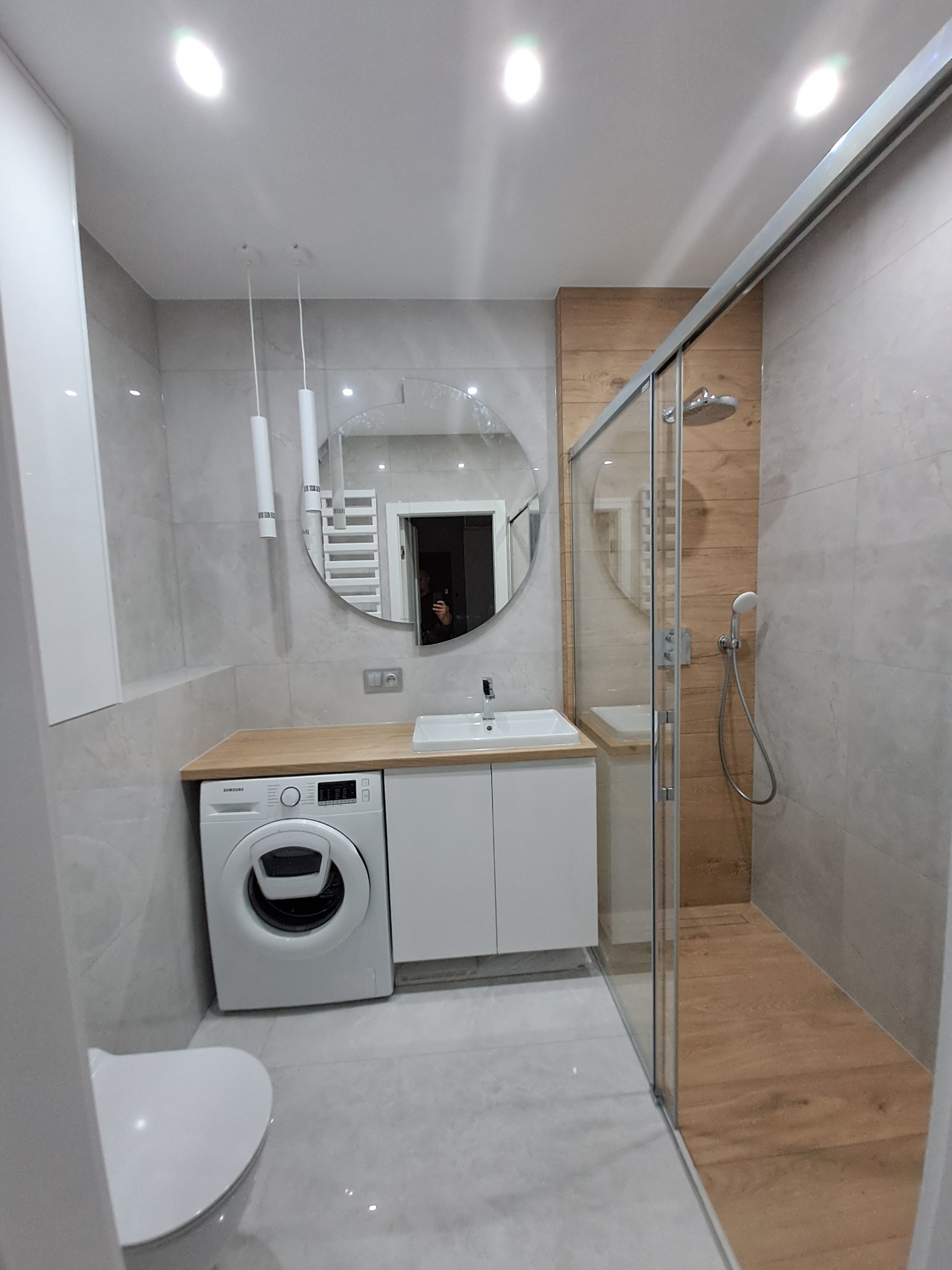 Nowoczesna łazienka z kabiną prysznicową typu walk-in, umywalką z szafką, pralką Samsung i okrągłym lustrem z podświetleniem LED, wykończona płytkami w odcieniach szarości i drewna.