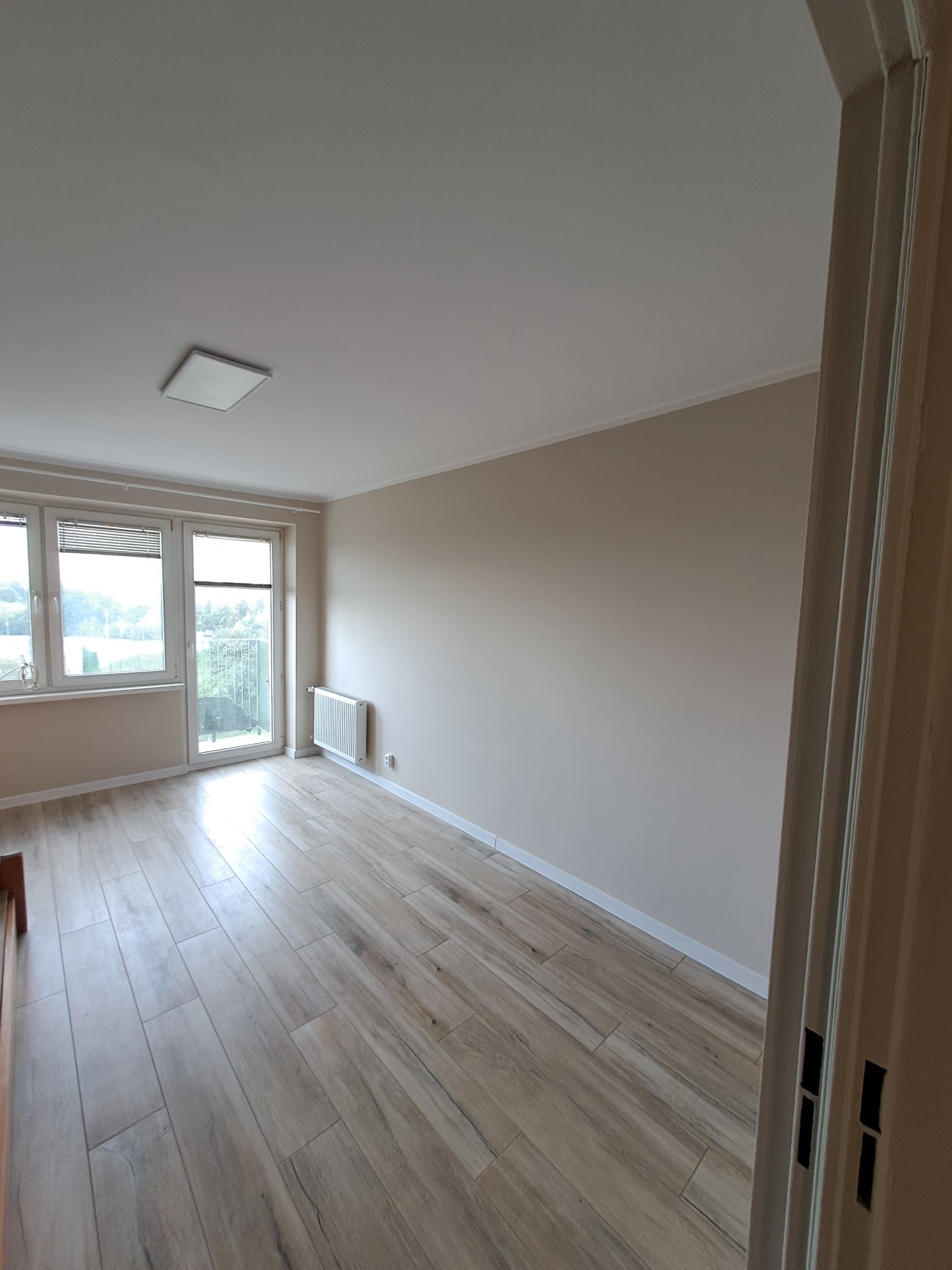 Wnętrze pustego pokoju z drewnopodobną podłogą z płytek, grzejnikiem pod oknem balkonowym i białymi ścianami, widok zza otwartych drzwi.