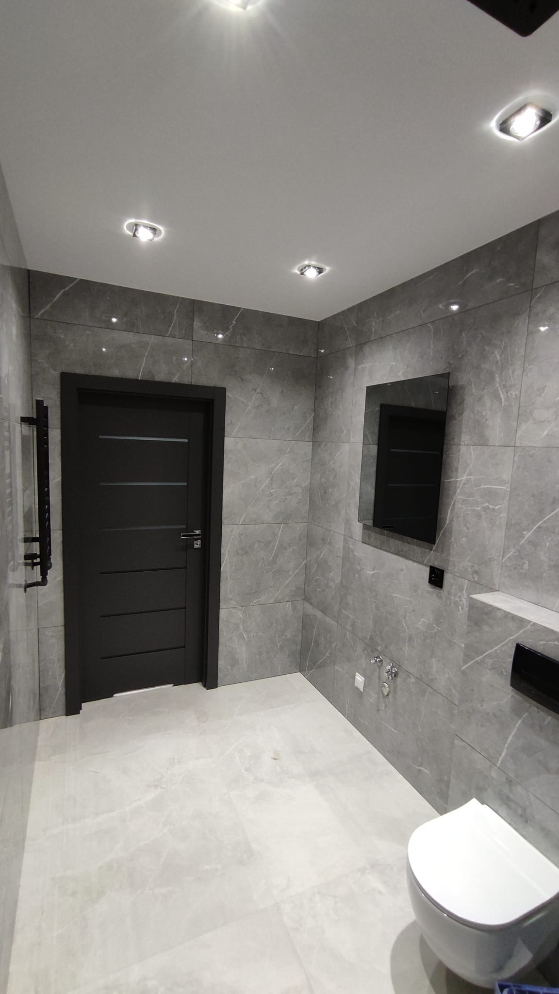 Nowoczesna łazienka z szarymi płytkami imitującymi marmur, czarnymi drzwiami z poziomymi frezowaniami, wiszącą toaletą i podświetlanym lustrem.