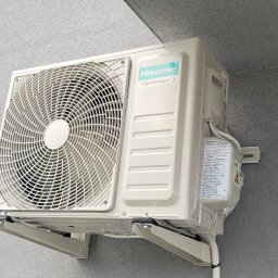 Otmech - Klimatyzator Hisense Inverter Expert zamontowany na szarej ścianie budynku, widoczny fragment okna z ciemną ramą.