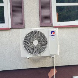 Otmech - Zamontowana jednostka zewnętrzna klimatyzacji marki Gree na elewacji budynku z brązowymi okiennicami i fragmentem zaniedbanego terenu z kostką brukową.