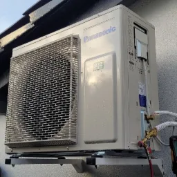 Zewnętrzna jednostka klimatyzacji Panasonic zamontowana na ścianie budynku, z podłączonym manometrem cyfrowym do pomiaru ciśnienia czynnika chłodniczego podczas instalacji.