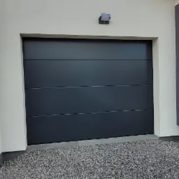 Nowoczesna, czarna segmentowa brama garażowa z poziomymi przetłoczeniami, zamontowana w jasnym budynku z szarą ścianą boczną i małym oknem, widok z zewnątrz na żwirowe podłoże.