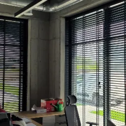Ciemne, poziome listwy w oknach biurowych, widok na zewnątrz częściowo zasłonięty, surowy betonowy filar i metalowe rury wentylacyjne w tle.