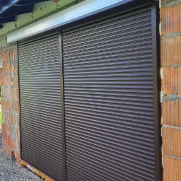 Zamknięte, brązowe rolety zewnętrzne zamontowane na surowej ścianie z cegły, widoczna piana montażowa i drewniany podest na żwirowym podłożu.