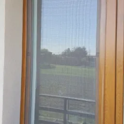 Okno z moskitierą w drewnianej ramie, widok na zewnątrz z zielenią i fragmentem balkonu.