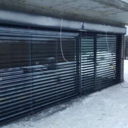 Zamknięte, ciemne żaluzje fasadowe zamontowane na budynku w zimowym krajobrazie, z widocznym śniegiem i drzewami w tle.