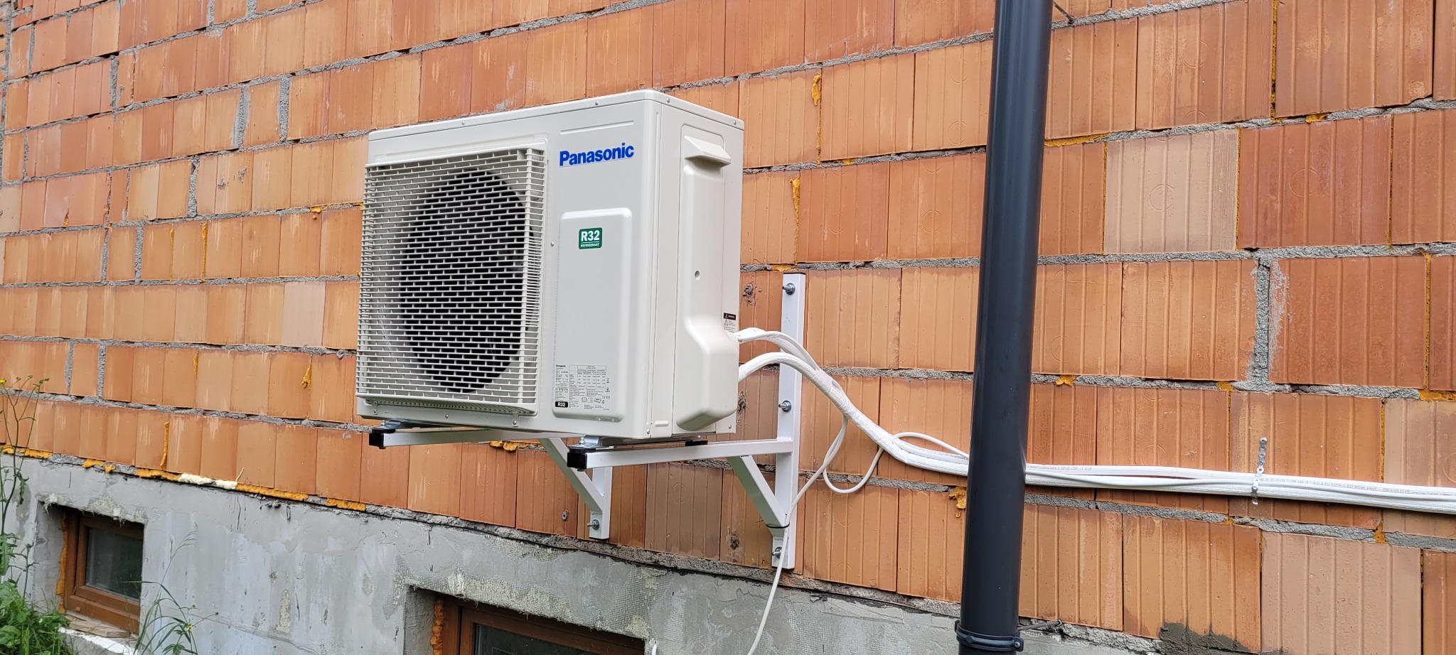 Zewnętrzna jednostka klimatyzacji Panasonic zamontowana na ścianie z cegły dziurawki, z widocznymi przewodami i rurą odprowadzającą skropliny.