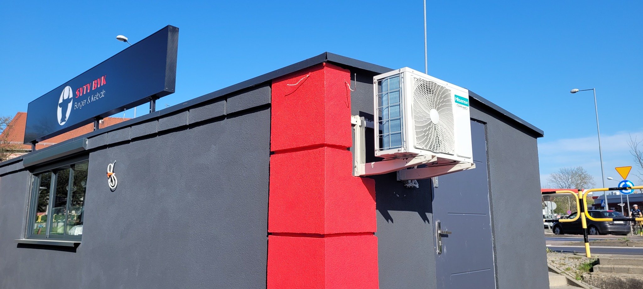 Zewnętrzny klimatyzator Hisense zamontowany na szarej ścianie budynku z czerwonym akcentem, widoczny szyld reklamowy i otoczenie miejskie.