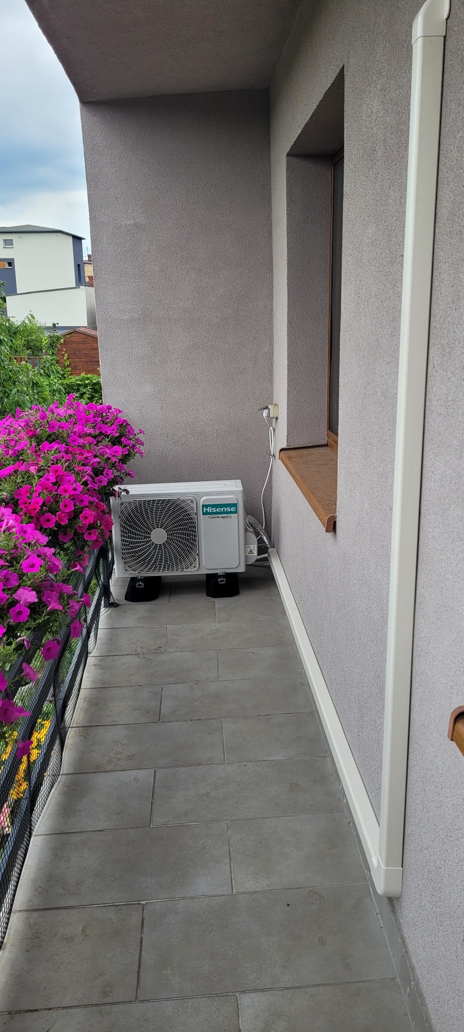 Biała jednostka zewnętrzna klimatyzacji Hisense zamontowana na balkonie z szarymi płytkami, z widoczną ścianą budynku o szarej, chropowatej strukturze i kwiatami w doniczce na balustradzie.