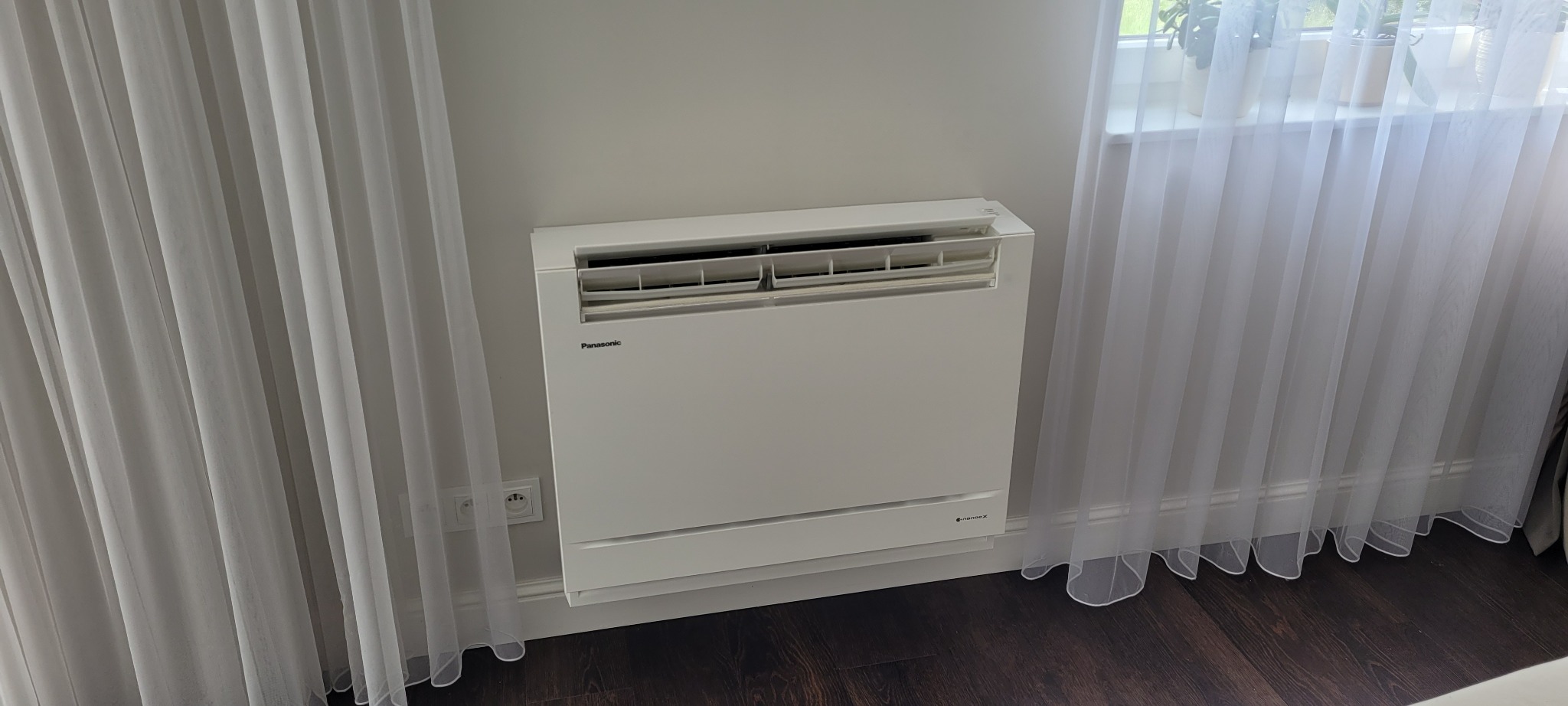 Biały, podłogowy klimatyzator Panasonic w nowoczesnym wnętrzu, widoczny fragment ściany, gniazdko elektryczne i zwiewne, białe zasłony.
