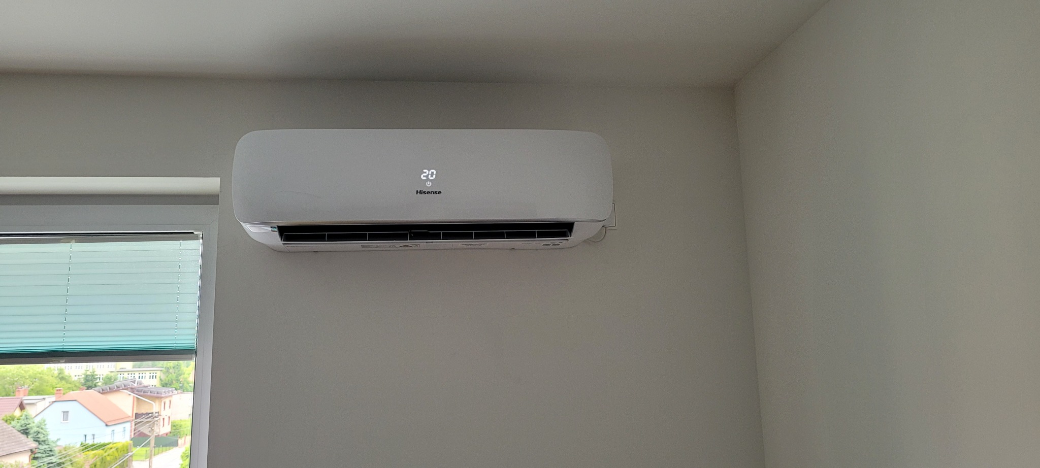 Biały klimatyzator Hisense zamontowany na ścianie w jasnym pokoju, widoczne okno z plisami i zabudowa domów na zewnątrz.