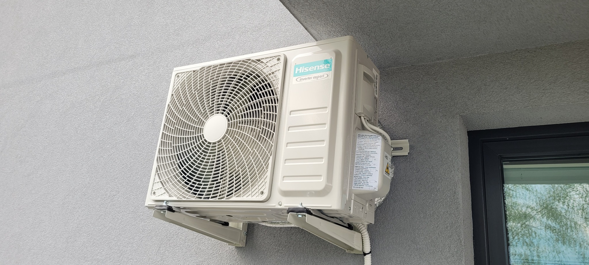 Klimatyzator Hisense Inverter Expert zamontowany na szarej ścianie budynku, widoczny fragment okna z ciemną ramą.