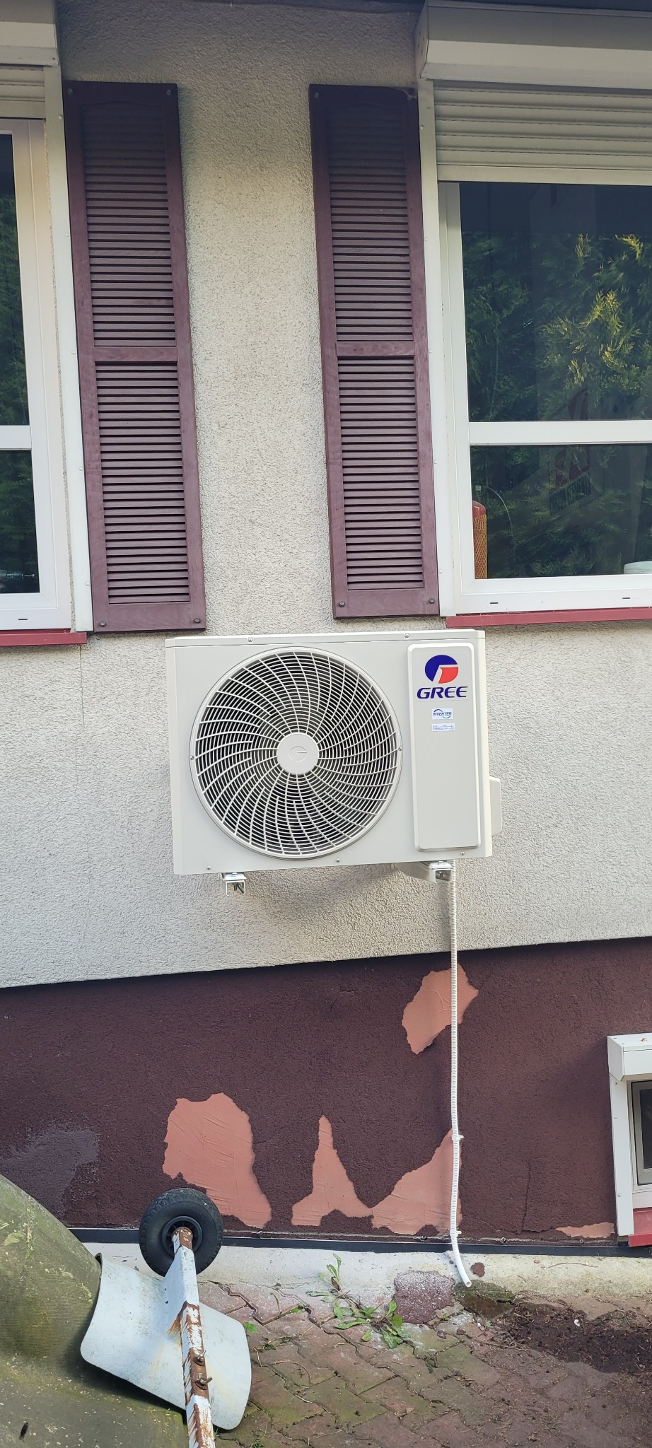 Zamontowana jednostka zewnętrzna klimatyzacji marki Gree na elewacji budynku z brązowymi okiennicami i fragmentem zaniedbanego terenu z kostką brukową.