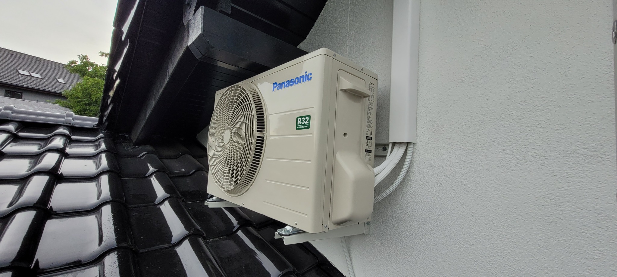 Zewnętrzna jednostka klimatyzacji Panasonic zamontowana na ścianie domu, widoczne rury odprowadzające, fragment czarnej dachówki w tle.