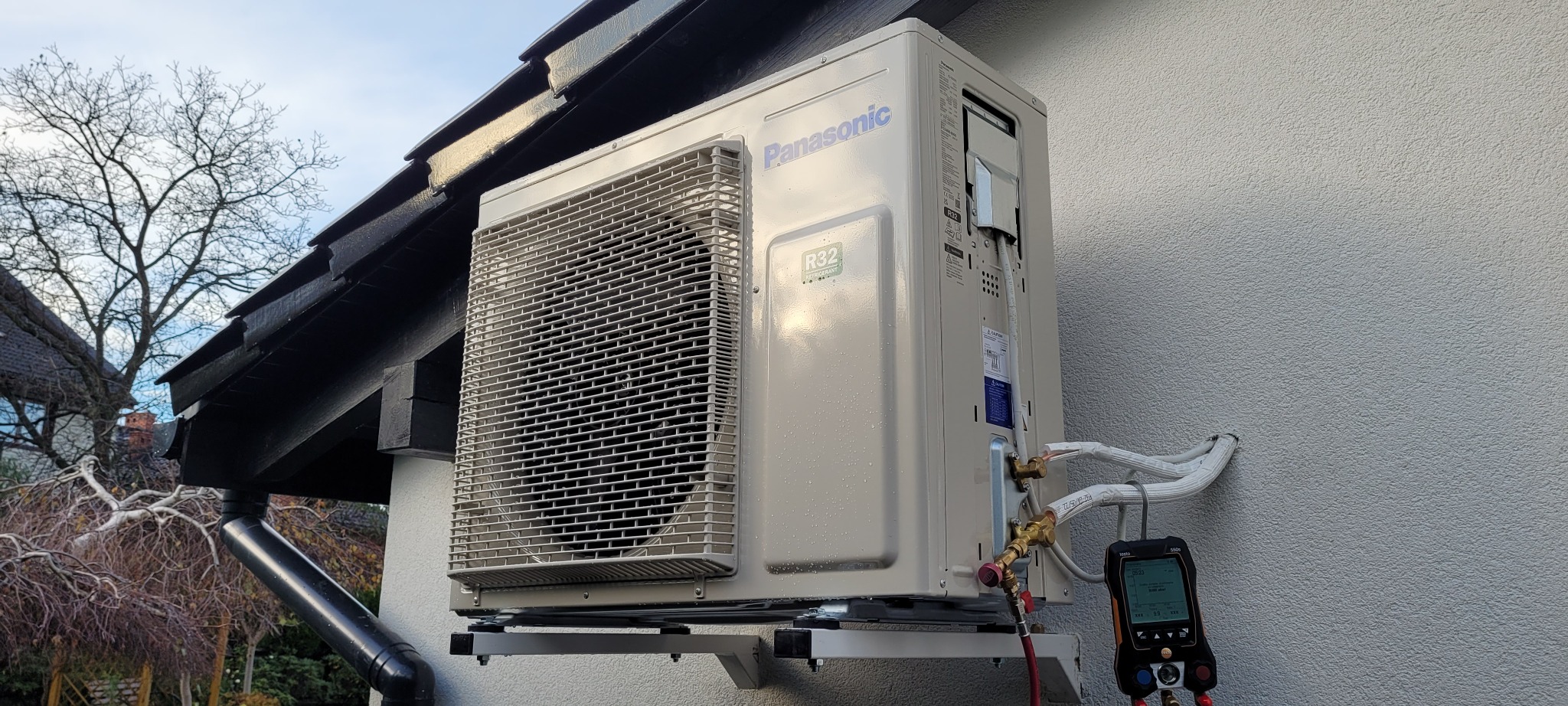 Zewnętrzna jednostka klimatyzacji Panasonic zamontowana na ścianie budynku, z podłączonym manometrem cyfrowym do pomiaru ciśnienia czynnika chłodniczego podczas instalacji.