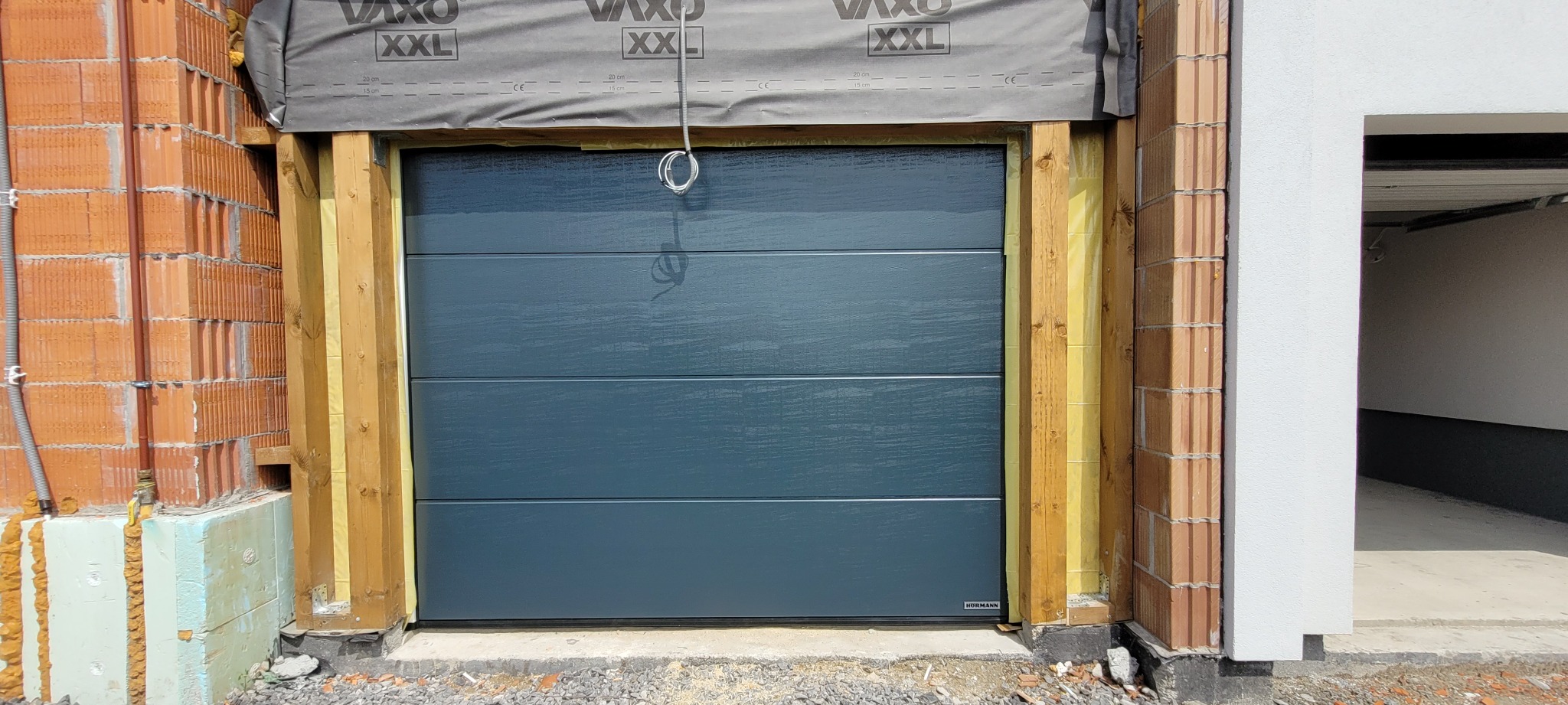 Montaż segmentowej bramy garażowej w surowym stanie, widoczne cegły, drewniana rama i izolacja, z zwisającym kablem instalacji elektrycznej.