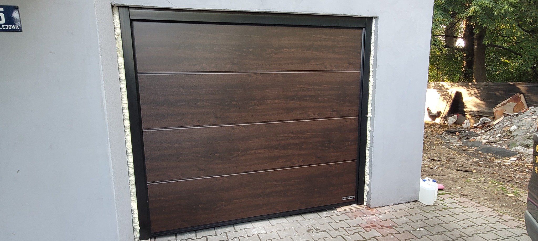 Zamknięta brama garażowa segmentowa z paneli imitujących drewno, czarna rama, widoczne pianowanie montażowe, w tle kury grzebiące w gruzach.
