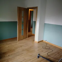 Wnętrze pokoju po remoncie z drewnopodobną podłogą, świeżo pomalowanymi ścianami w kolorze białym i turkusowym oraz otwartymi drewnianymi drzwiami z przeszkleniami. Na podłodze leży wąż od odkurzacza.