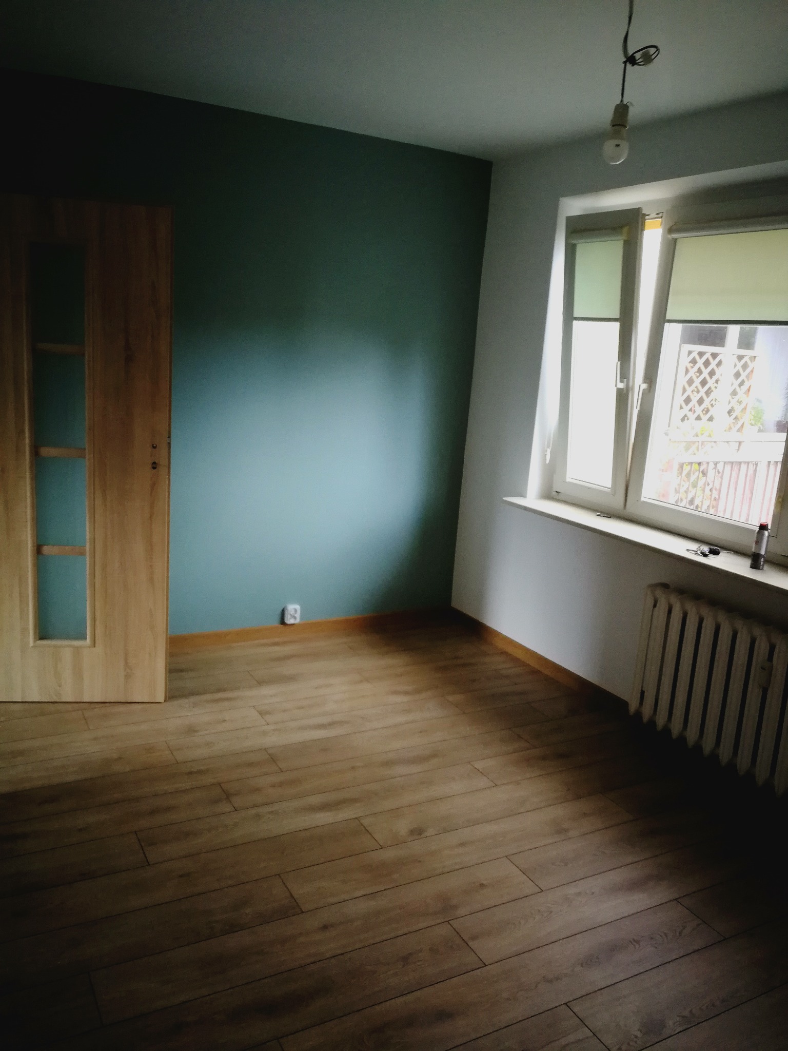 Pusty pokój po renowacji z drewnianą podłogą, pomalowaną na niebiesko ścianą i białym oknem z roletą, widoczny grzejnik i wisząca żarówka.