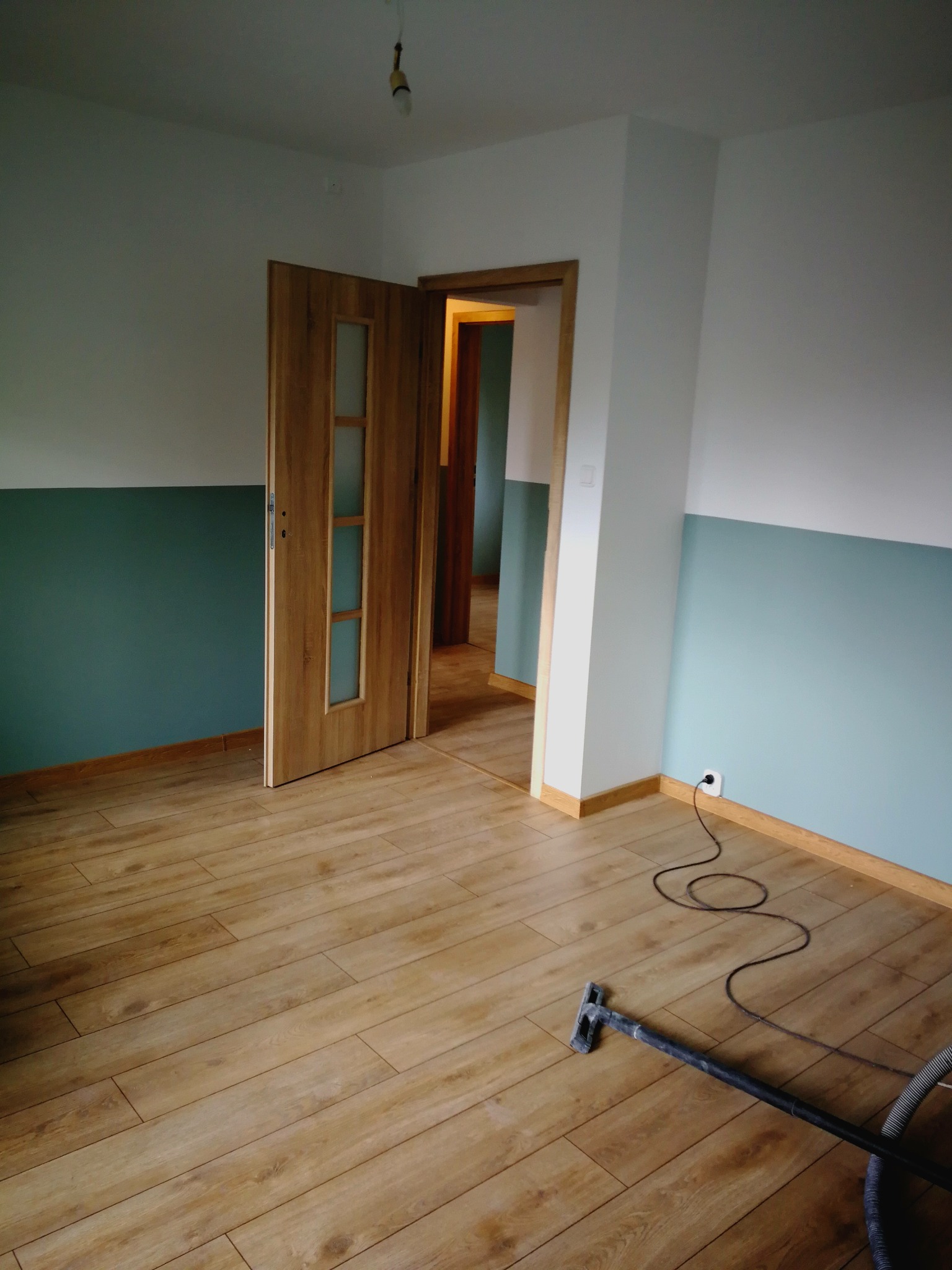Wnętrze pokoju po remoncie z drewnopodobną podłogą, świeżo pomalowanymi ścianami w kolorze białym i turkusowym oraz otwartymi drewnianymi drzwiami z przeszkleniami. Na podłodze leży wąż od odkurzacza.